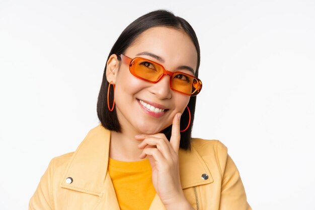 Nahaufnahme des Porträts einer asiatischen Frau, die denkt, eine Sonnenbrille zu tragen und lächelnd nach oben zu schauen, nachdem sie über weißem Studiohintergrund steht