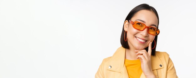 Nahaufnahme des Porträts einer asiatischen Frau, die denkt, eine Sonnenbrille zu tragen und lächelnd nach oben zu schauen, nachdem sie über weißem Studiohintergrund steht