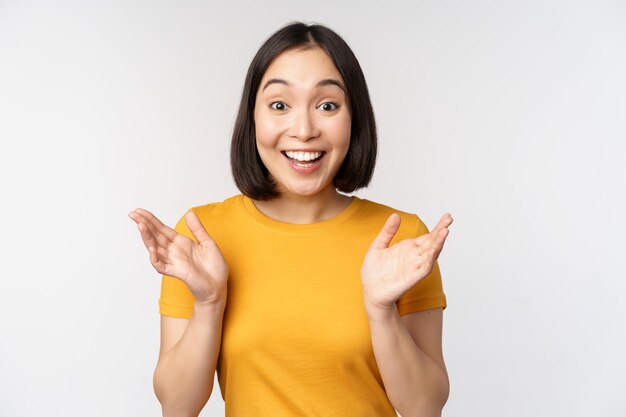 Nahaufnahme des Porträts einer asiatischen Frau, die überrascht aussieht, wow Gesicht beeindruckt in die Kamera starrt, die über weißem Hintergrund in gelbem T-Shirt steht