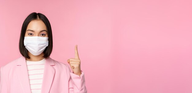 Nahaufnahme des Porträts einer asiatischen Geschäftsfrau in medizinischer Gesichtsmaske und Anzug, die mit dem Finger nach oben zeigt und das obere Werbebanner zeigt, das über rosa Hintergrund steht