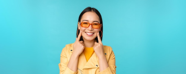 Nahaufnahme des Porträts einer asiatischen jungen Frau mit Sonnenbrille, die lächelt und romantisch aussieht und glücklich über blauem Hintergrund steht