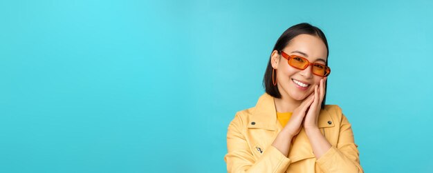 Nahaufnahme des Porträts einer asiatischen jungen Frau mit Sonnenbrille, die lächelt und romantisch aussieht und glücklich über blauem Hintergrund steht