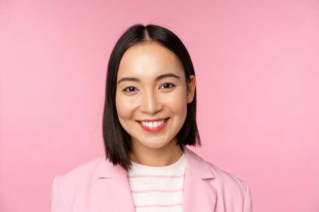 Nahaufnahme des Porträts einer asiatischen Unternehmensfrau, die professionell in die Kamera lächelt und einen Anzug trägt, der über rosafarbenem Hintergrund steht Kopierraum