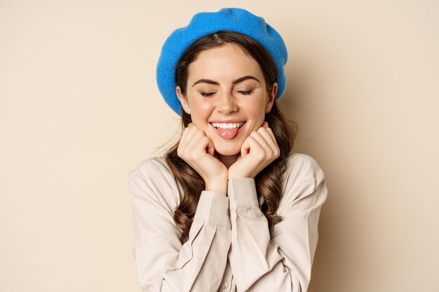 Nahaufnahme des Porträts einer femininen jungen Frau mit trendigem französischem Hut, die süß und romantisch posiert, ein albernes kokettes Gesicht und ein zartes Lächeln macht, beiger Hintergrund.