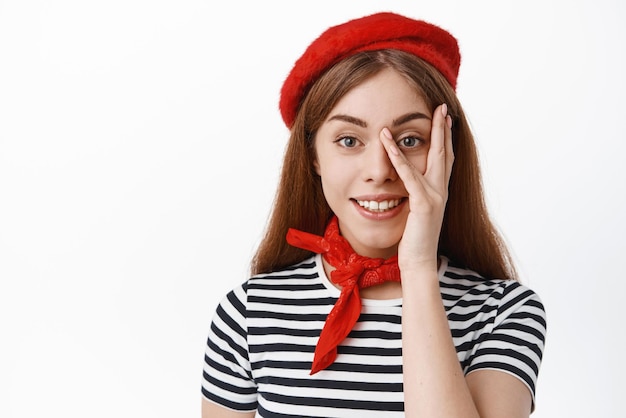 Nahaufnahme des Porträts einer jungen Studentin in französischer Baskenmütze, die die Hände auf dem Gesicht hält und die saubere Gesichtshaut mit natürlichem Make-up berührt, das auf den weißen Hintergrund der Kamera lächelt