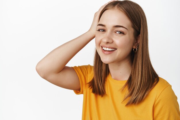 Nahaufnahme des Porträts einer schönen, aufrichtigen Frau, die glücklich lächelt, die Frisur berührt und sorglos nach vorne schaut, in gelbem T-Shirt gegen weiße Wand stehend