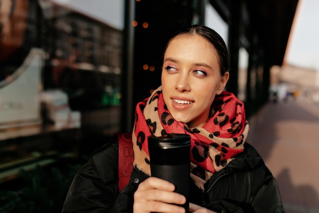 Nahaufnahme des Porträts einer schönen jungen Frau mit hellem Make-up, die einen roten Schal hält, der Kaffee hält und im Sonnenlicht in der Stadt spazieren geht Grünäugige Brünette, die Freizeit in der Stadt genießt