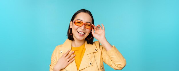 Nahaufnahme des Porträts einer stilvollen asiatischen Frau mit Sonnenbrille, die lacht und lächelt und glücklich aussieht, wenn sie in trendiger Kleidung auf blauem Hintergrund posiert