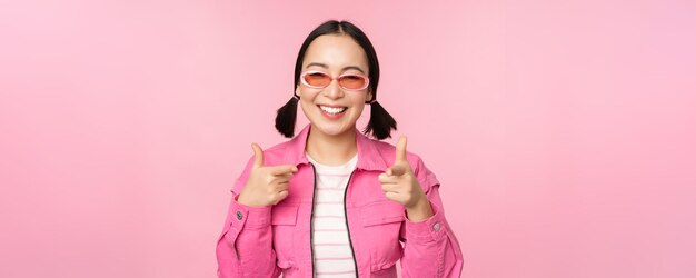 Nahaufnahme des Porträts eines modernen asiatischen Mädchens mit Sonnenbrille, das mit dem Finger auf die Kamera zeigt und Sie einlädt oder beglückwünscht, wenn Sie über rosa Hintergrund stehen