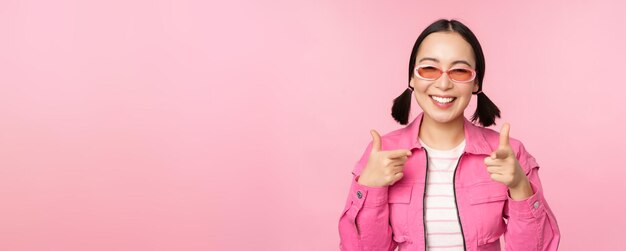 Nahaufnahme des Porträts eines modernen asiatischen Mädchens mit Sonnenbrille, das mit dem Finger auf die Kamera zeigt und Sie einlädt oder beglückwünscht, wenn Sie über rosa Hintergrund stehen