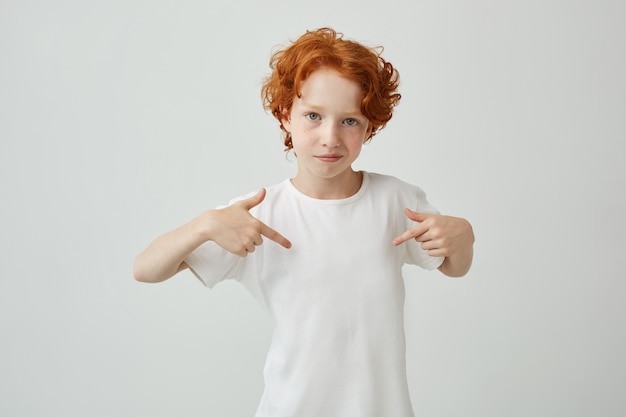 Nahaufnahme des rothaarigen niedlichen Jungen mit Sommersprossen, die mit den Fingern auf weißem T-Shirt mit ernstem und selbstbewusstem Ausdruck zeigen. Speicherplatz kopieren.