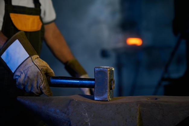 Nahaufnahme des schmieds in schutzhandschuhen mit schwerem hammer für arbeiten, die metall auf amboss schlagen manuelle und harte arbeit in der schmiede fertigungskonzept