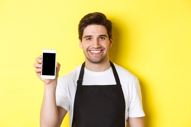 Nahaufnahme des schönen Kellners in der schwarzen Schürze, die Smartphonebildschirm zeigt, App empfehlend, über gelbem Hintergrund stehend.