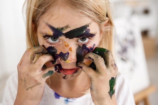 Nahaufnahme des schönen kleinen europäischen Mädchens mit blonden Haaren und Sommersprossen, das mit ihren großen Augen in die Kamera schaut und ihr Gesicht mit den Händen in Farbe berührt