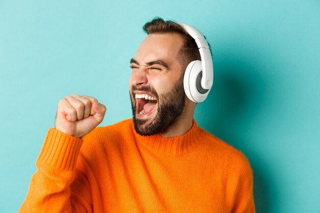 Nahaufnahme des schönen modernen Mannes, der Musik in den Kopfhörern hört, im orange Pullover stehend
