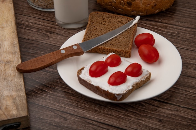 Nahaufnahme des Tellers der Roggenbrotscheiben, die mit Hüttenkäse und Tomaten und Messer auf hölzernem Hintergrund verschmiert werden