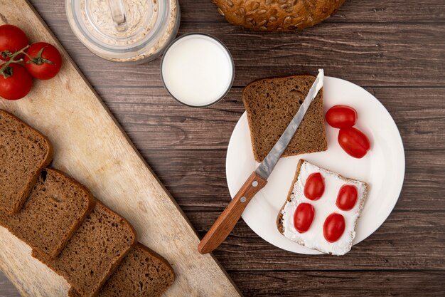 Nahaufnahme des Tellers mit Roggenbrotscheiben und Tomaten und Messer mit Milchhaferflocken auf hölzernem Hintergrund