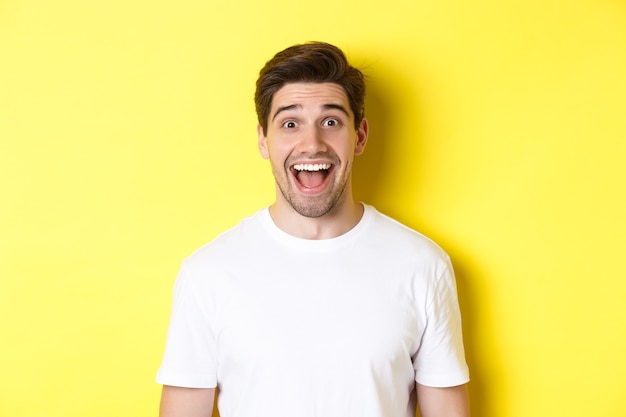Nahaufnahme des überraschten gutaussehenden Kerls, der auf gute Nachrichten reagiert und über gelbem Hintergrund im weißen T-Shirt steht.