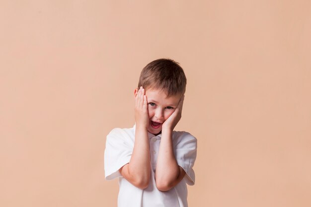 Nahaufnahme des überraschten netten Jungen mit dem offenen Mund, der nahe Beige steht, färbte Wand