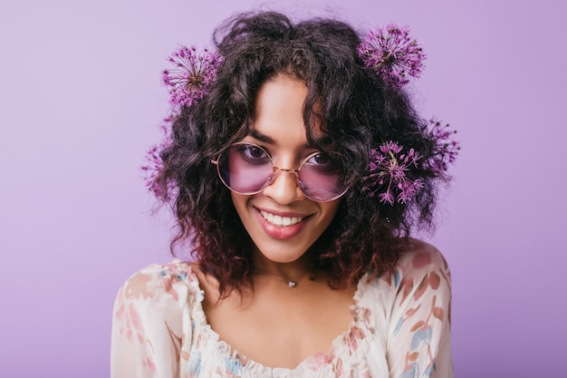 Nahaufnahme des wundervollen weiblichen Modells in der stilvollen Sonnenbrille, die herumalbert. Innenporträt des gewinnenden afrikanischen Mädchens mit lila Blumen im schwarzen Haar.