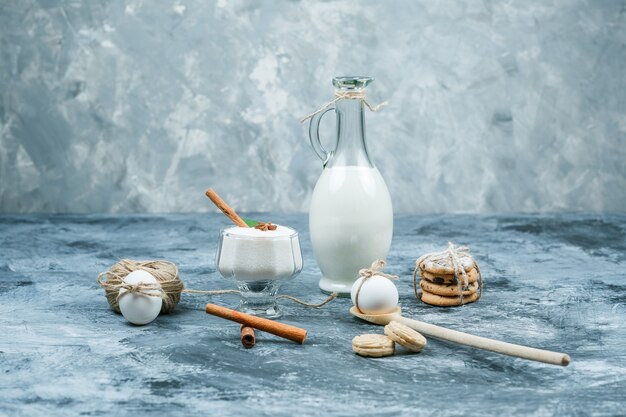 Nahaufnahme ein Krug Milch und eine Glasschale Joghurt mit Keksen, Eiern, Clew und Zimt auf dunkelblauer und grauer Marmoroberfläche. horizontal