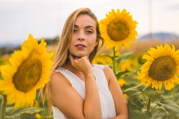 Nahaufnahme einer Blondine mit einem weißen Kleid, die in einem Sonnenblumenfeld posiert