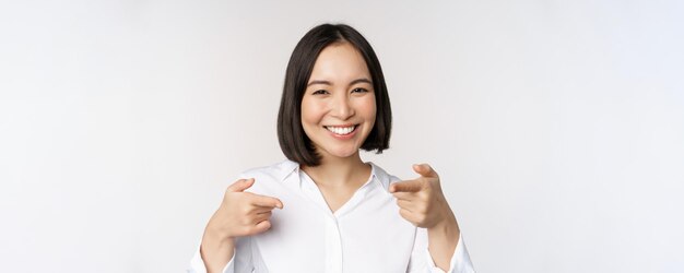 Nahaufnahme einer enthusiastischen jungen Frau, die lächelt und mit dem Finger auf die Kamera zeigt und Sie auswählt, einladende und rekrutierende Menschen zu gewinnen, die über weißem Hintergrund stehen