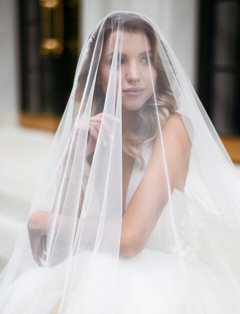 Nahaufnahme einer ernsthaften und attraktiven Frau in einem langen weißen Kleid, die die Hand hebt, das Gesicht unter dem Schleier versteckt und wegschaut, während sie auf dem Hintergrund eines modernen Gebäudes sitzt