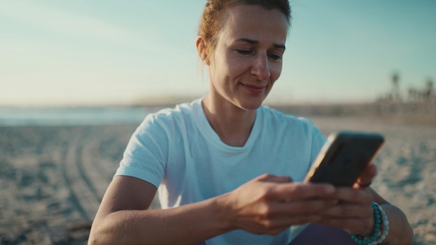 Nahaufnahme einer Frau, die ihre sozialen Medien mit dem Smartphone am Meer überprüft Sportliches Mädchen, das sich nach dem Training am Strand ausruht