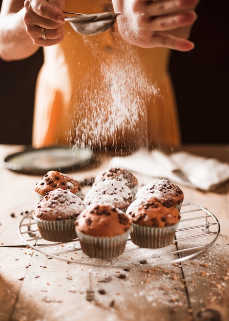 Nahaufnahme einer Frau, die Puderzucker auf Muffins über dem abkühlenden Gestell staubwischt