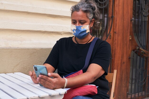 Nahaufnahme einer Frau mit Gesichtsmaske, während sie ihr Telefon auf dem Tisch im Freien eines Cafés benutzt