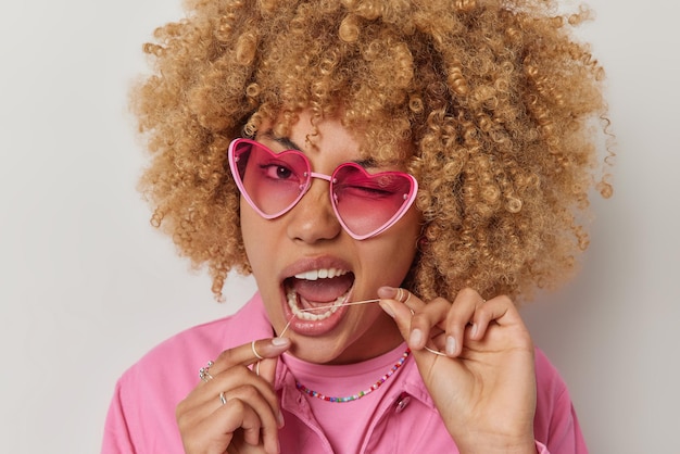 Kostenloses Foto nahaufnahme einer frau mit lockigem haar zwinkert, trägt eine rosafarbene sonnenbrille und eine jacke verwendet zahnseide zum reinigen der zähne. entfernt lebensmittel, die über grauem studiohintergrund isoliert sind