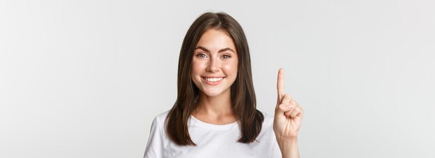 Nahaufnahme einer fröhlichen, attraktiven jungen Frau, die lächelnd einen weißen Hintergrund mit einem Finger zeigt