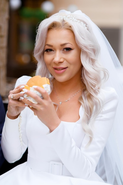 Nahaufnahme einer fröhlichen und hübschen Braut mit Frisur und Accessoires auf dem Kopf, die ein Design-Hochzeitskleid mit langen Ärmeln und Gürtel an der Taille trägt und ein Sandwich hält, während sie im Freien in die Kamera schaut