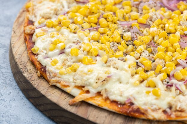 Nahaufnahme einer ganzen leckeren Pizza mit geschmolzenem Käse, Mais und Zwiebeln auf einem Brett