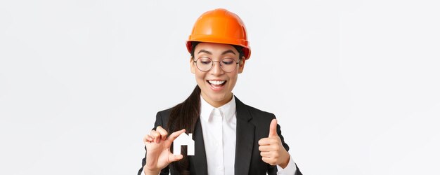 Nahaufnahme einer glücklichen, erfreuten asiatischen Architektin, Bauingenieurin in Helm und Anzug, die Miniatur zu Hause betrachtet und Daumen hoch zeigt, garantiert die Qualität der Reparaturarbeiten auf weißem Hintergrund