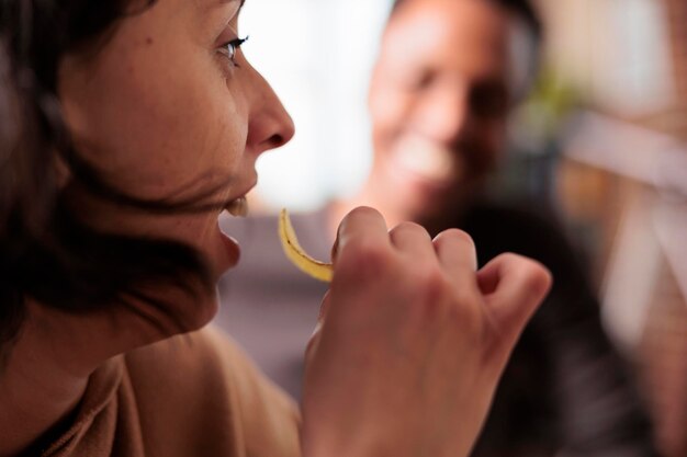 Nahaufnahme einer glücklichen Frau, die Chips isst, während sie zu Hause mit multiethnischen Freunden sitzt. Fröhliche Person, die Snacks genießt, während sie mit verschiedenen Menschen am Tisch im Wohnzimmer sitzt.