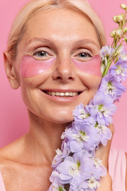 Nahaufnahme einer glücklichen Frau mit gesunder, sauberer Haut, die sanft lächelt und feuchtigkeitsspendende Flecken unter den Augen aufträgt, hält Blumen in der Nähe von Gesichtsposen vor rosafarbenem Hintergrund. Gesichtspflege und Schönheitskonzept.