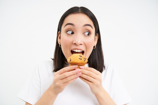 Nahaufnahme einer glücklichen koreanischen Frau, die ein köstliches Cupcake-Dessert isst, lächelnd vor weißem Hintergrund