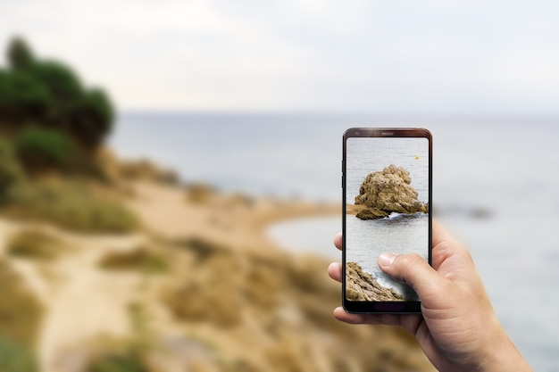 Nahaufnahme einer Hand, die ein Telefon hält und ein Foto von einem Strand macht