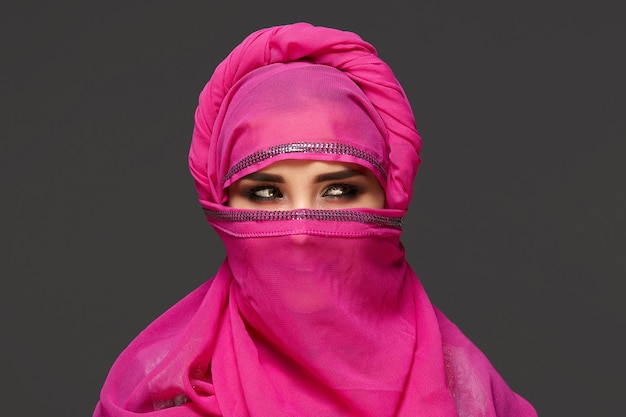 Nahaufnahme einer jungen Frau mit hellem Make-up, die den rosa Hijab trägt, der mit Pailletten verziert ist. Sie posiert im Studio und sieht schlau auf einem dunklen Hintergrund aus. Menschliche Emotionen, Gesichtsausdruck