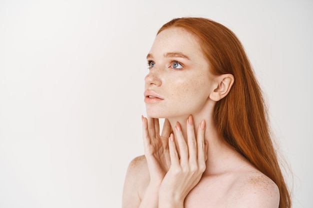 Nahaufnahme einer jungen Frau mit langen roten Haaren, die im Profil steht und mit blauen Augen aufschaut, nackt mit blasser, glatter Haut stehend