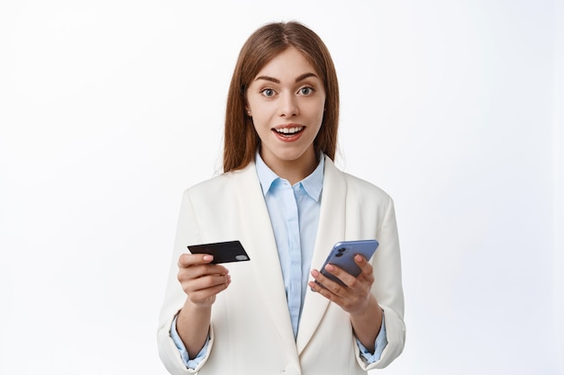 Kostenloses Foto nahaufnahme einer jungen geschäftsfrau im anzug, die einfach online mit plastik-kreditkarte und smartphone bezahlt, online einkaufen, handy halten, erstaunt auf die vorderseite schauen, weiße wand