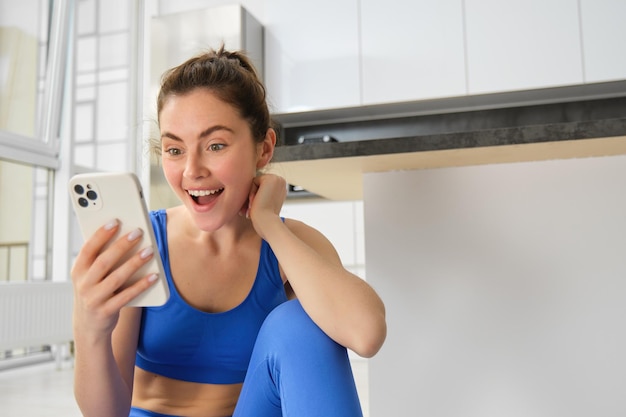 Kostenloses Foto nahaufnahme einer jungen, glücklichen frau, die zu hause sport trainiert, ihr handy überprüft und erstaunt sieht