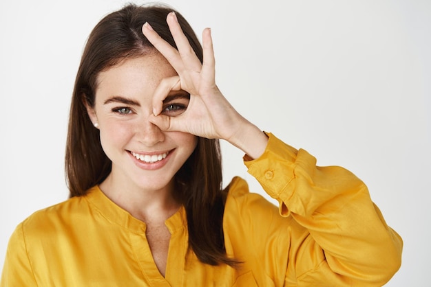 Nahaufnahme einer jungen selbstbewussten Frau, die ein okayzeichen auf dem Auge zeigt, lächelnd und durchsetzungsfähig aussieht und Ja sagt oder die Zustimmung erteilt, die über weißem Hintergrund steht