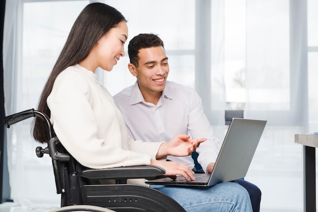 Nahaufnahme einer lächelnden jungen Frau, die auf dem Rollstuhl zeigt zu ihrem Kollegen auf Laptop sitzt