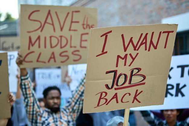 Nahaufnahme einer Menschenmenge, die bei öffentlichen Demonstrationen gegen Arbeitslosigkeit protestiert. Der Fokus liegt auf dem Banner mit der Aufschrift "Ich will meinen Job zurück".