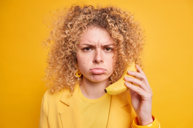 Nahaufnahme einer mürrischen, lockigen Frau, die Banane hält, während das Telefon in der Nähe des Ohrs vorgibt, jemanden anzurufen, der einen unzufriedenen Ausdruck in gelber Kleidung hat, drückt negative Emotionen aus