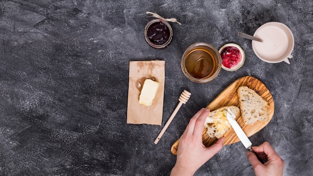 Nahaufnahme einer Person, die Butter mit Messer hinzufügt; Himbeermarmelade und Honig auf schwarzem strukturiertem Hintergrund