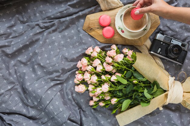 Nahaufnahme einer Person, die Makrone im Kaffee mit Kamera- und Blumenblumenstrauß auf Tischdecke eintaucht